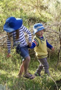 Blonde girl wearing a blue brad-brimmed hat, white long sleeved shirt with blue horizontal stripes, denim shorts holds the hand of a smaller boy, wearing a blue hat, blue long-sleeved shirt, yellow sweater vest and beige trousers. Both children wear sneakers as they walk through grasslands and bush.