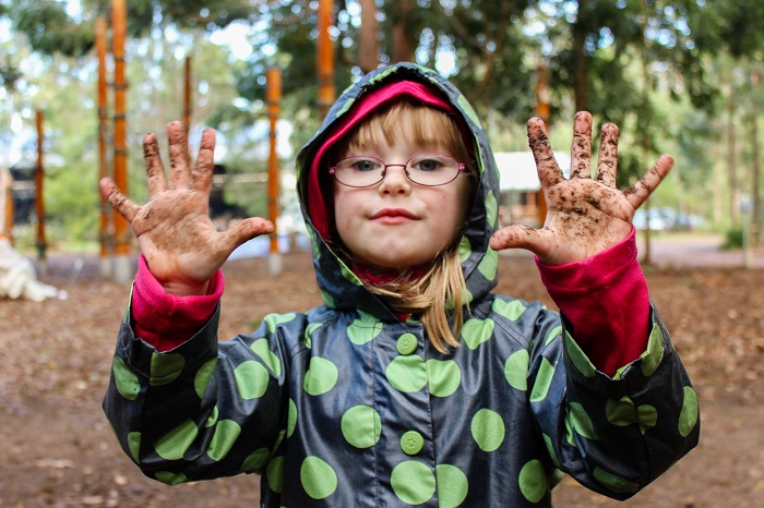 girl wearing glasses and green spotted raincoat with muddy hands