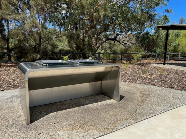 This is one of the universal barbecue areas at Kent Street Weir. The stainless steel barbecue is silver in colour, and sits on a concrete slab. The barbecue sits in an open space, surrounded by woodchips with trees in the background. The trees are brown with green leaves.