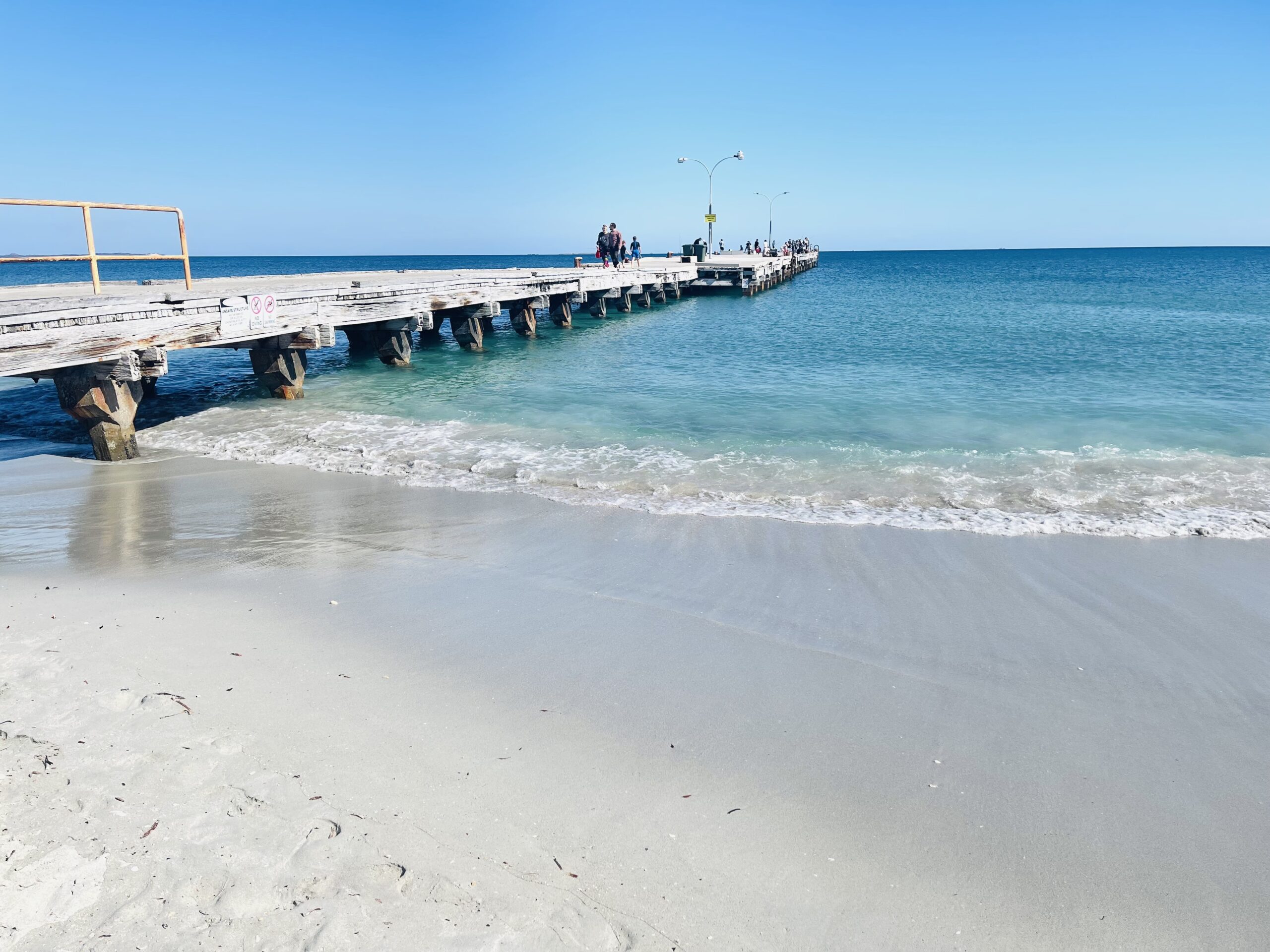This photograph was taken on the shore of Woodman Point Beach overlooking Ammo Jetty. Ammo Jetty is an old wooden bridge, which has faded to a light brown colour over many years of use. There are people on the bridge in the distance. It is a bright and sunny day with no clouds, white sand and clear light blue water.