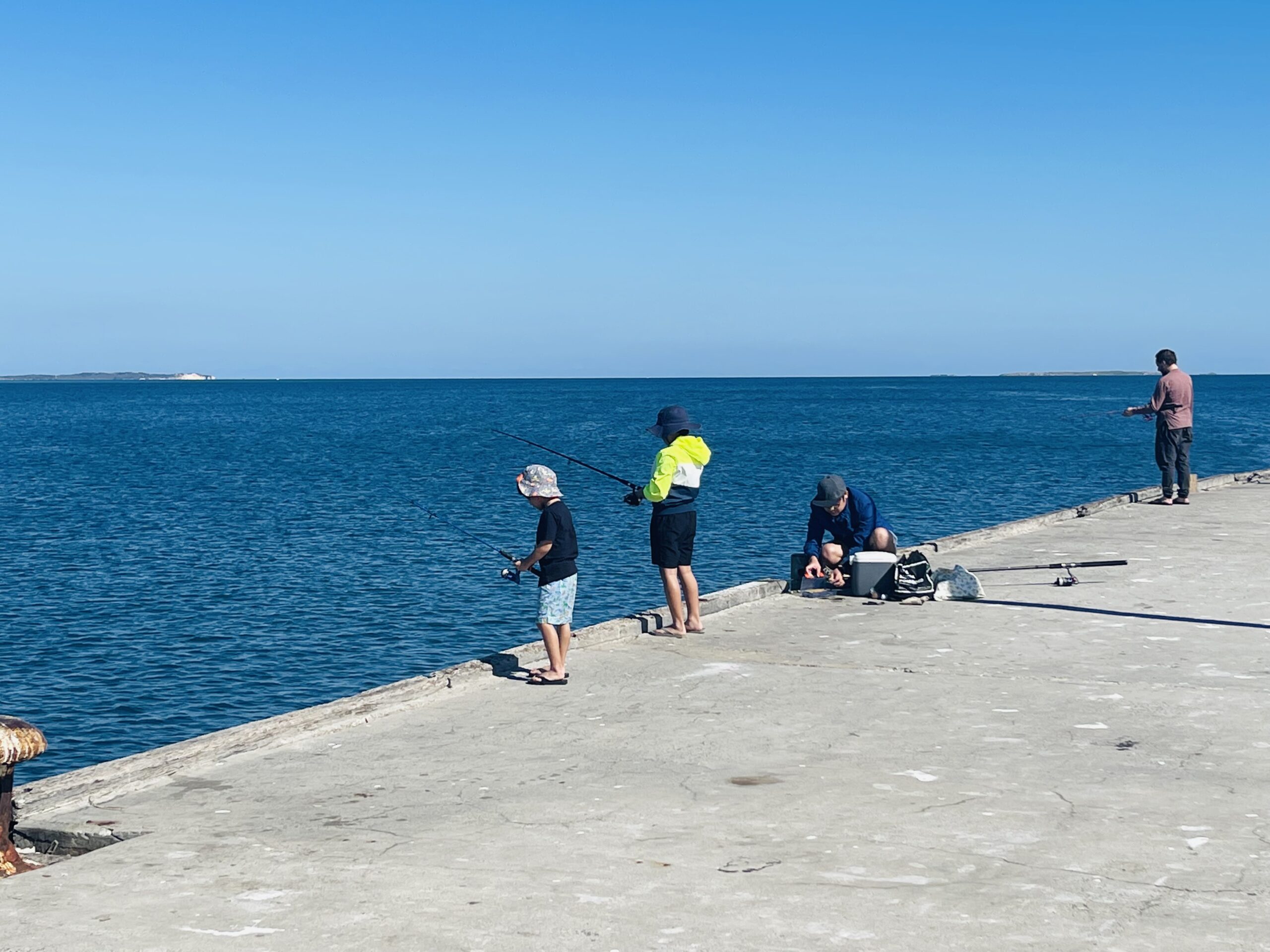 This photo was taken on the Ammo Jetty and shows two children and two adults fishing off the side of the jetty. The pathway is concrete and light silver in colour. The ocean is a deep blue, with the sky above a baby blue colour.