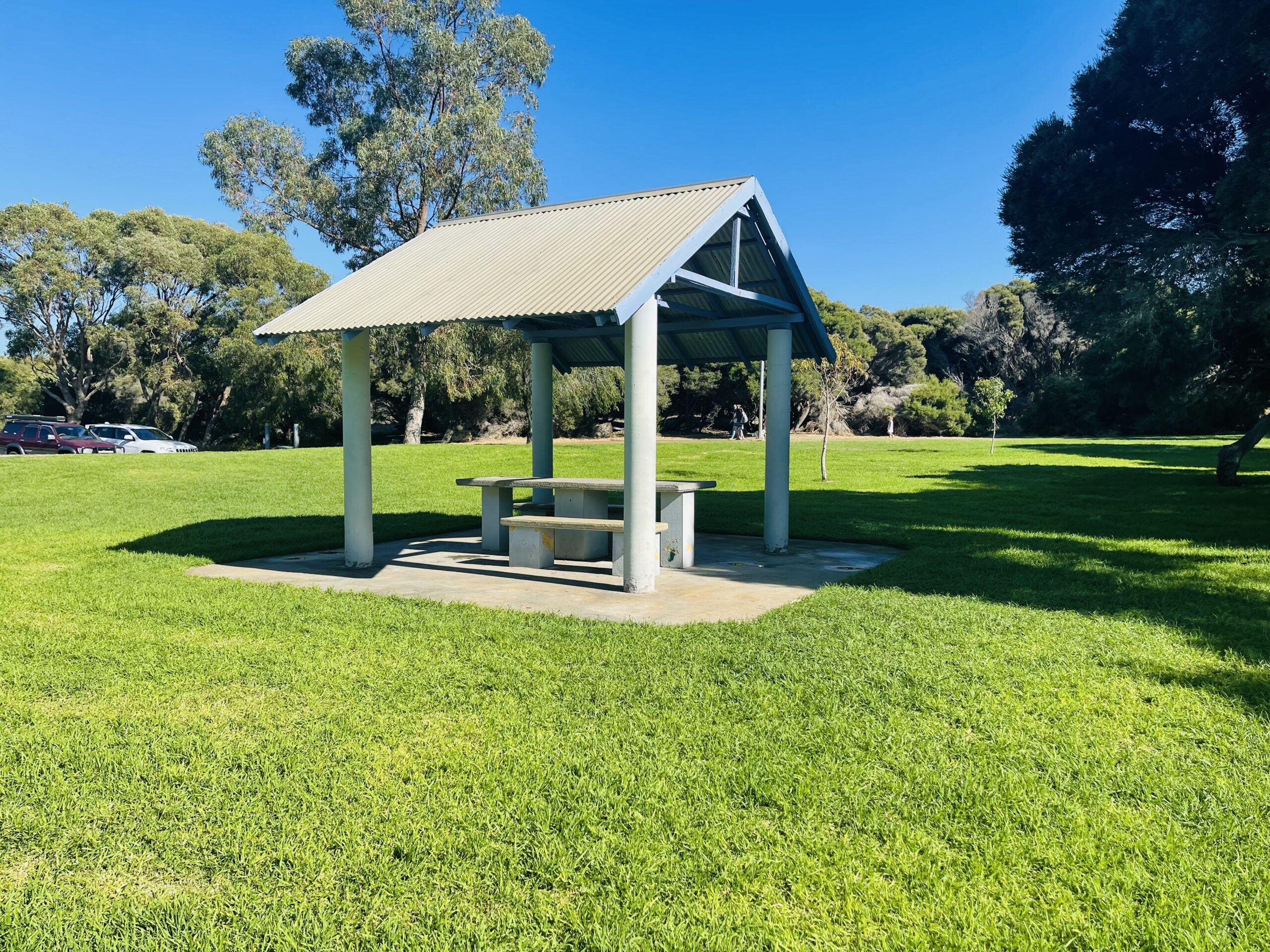 This photograph shows the closest sheltered picnic table to the barbecue. This structure has four tall grey beams and a grey tin roof with blue beams. The picnic table is concrete and grey in colour, and sits on a grey concrete slab.