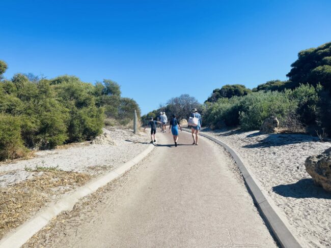 This photograph was taken of a family walking away from the camera, towards Woodman Point Beach. The family are following the open pathway which is concrete and surrounded by white sand and green shrubbery.