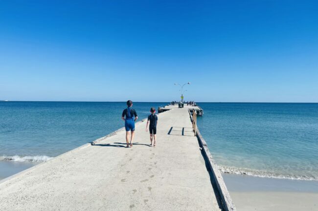 This photo was taken with Ammo Jetty being the central focus. The Jetty extends out across the water, with two kids walking away from the camera into the distance. The jetty has a light grey concrete pathway, with brown wooden framing on each side. The sand on the beach is white, which extends to the clear blue water. The sky above is blue with no clouds in sight. 