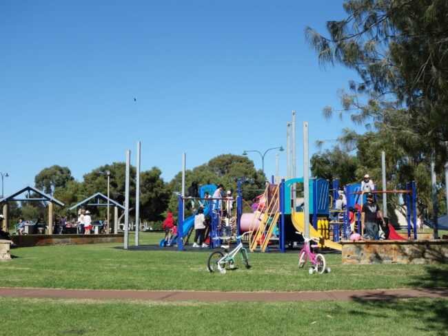 This image was taken of the playground at John Graham Reserve. The playground is bright in colour and features swings, slides and tunnels. People are using the playground in the distance, and there are two children's bicycles in the centre of the image. The playground is surrounded by green grass, green trees and a bright blue sky.