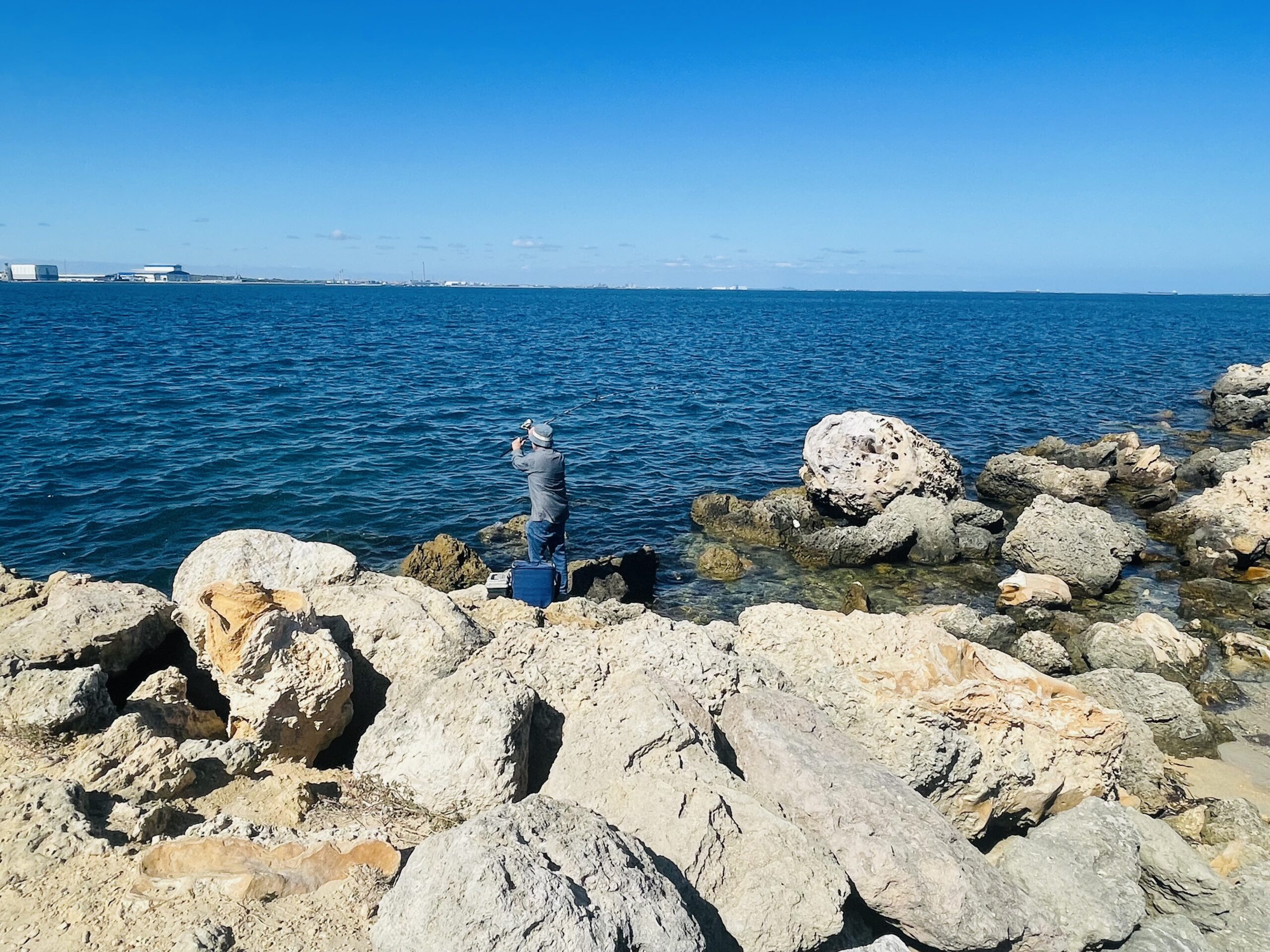 This photograph was taken of a man rock fishing at Woodman Point Headland. The man is standing on the cream-coloured rocks as he lifts his fishing rod over his head to cast into the dark blue ocean
