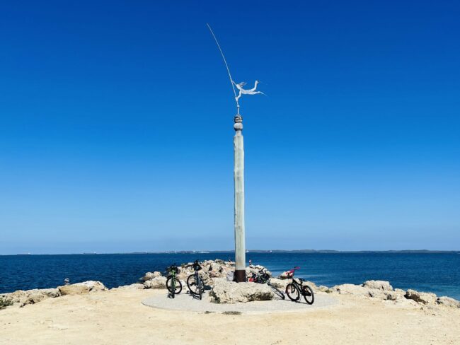 This photograph was taken of the sculpture located at Woodman Point Headland. The sculpture is steel and grey, and sits on a tall, light coloured wooden pole. There are four bicycles resting at the bottom of the sculpture. There are dirt paths surrounding the sculpture which sits on a bed of rocks overlooking the blue ocean. The sky is blue and there are no clouds in the sky.