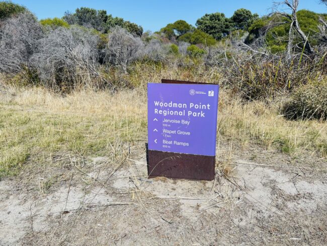 This is a photo of a large purple sign on top of a brown metal sculpture. Text on the sign reads “Woodman Point Regional Park. Jervoise Bay, 500m. Wapet Grove, 1.7km, Boat Ramps 600m.”. The sign features the logo of the Department of Parks and Wildlife.