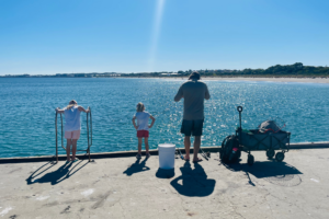 A father and two young girls stand on the edge of the Woodman Point Ammo Jetty on a sunny day. They are looking down into the crystal blue water, which can be seen in the midground of the picture, and are facing away from the camera. They have a wagon, fishing rods and bucket of bait beside them. The sky is bright blue and clear.