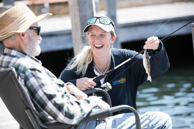 A volunteer from Fishability assists an older man with fishing on the Woodman Point Ammo Jetty. She is crouched down next to the man who is sitting on a camping chair. She has wears a beige coloured hat with black sunglasses sitting on the peak and a long sleeve blue shirt with the Fishability logo on the right. Her blonde hair is in a pony tail and sits on her shoulder. She is holding a fishing rod and a fish that has been caught, and is grinning at the older man. The older man grins back at her. He is wearing a yellow, wide-brim sun hat, black sunglasses and a checkered long-sleeve button-up shirt that is green, grey and white in colour with light blue jeans. The water can just be seen behind them, but it is out of focus.