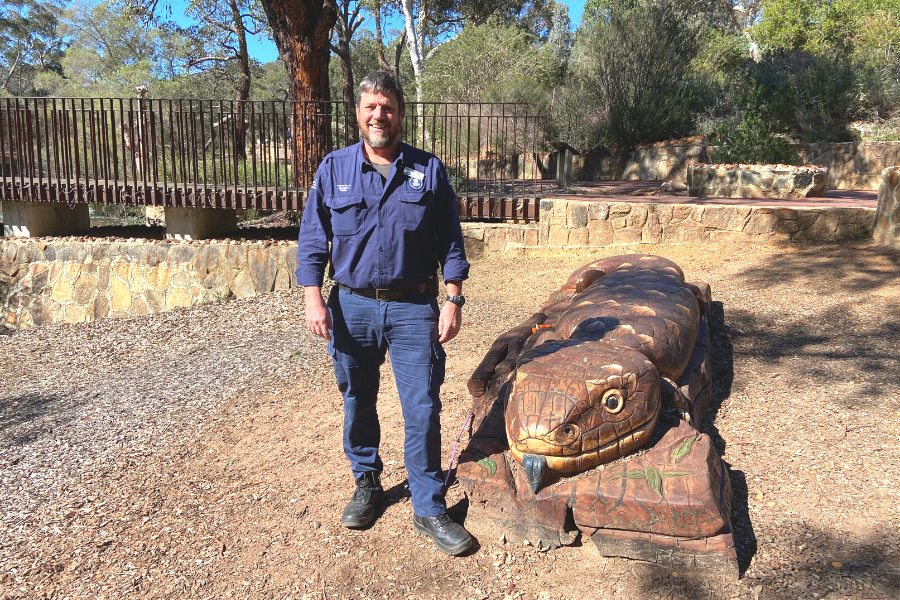 A photograph of Paul Udinga. He is standing in the nature playground at John Forrest National Park, next to a wooden sculpture of a lizard. He is smiling at the camera and is wearing his navy blue ranger uniform. The ground is covered in brown dirt and woodchips, and the background features bush and trees, small limestone walls and a rust-red steel bridge.