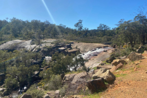 A photograph of National Park Falls at John Forrest National Park. The photo has been taken from the side, and quite a distance away. There are lots of brown-grey rocks and sheets of granite, as well as green bushes and trees. The waterfall is flowing over a softly slanted sheet of granite. The sky is bright blue.