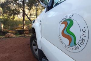 A photo of the side of a car parked at Mundy Regional Park. Only the left, front-half of the white car is visible. On the passenger door is a large sticker, which is the logo of the Friends of Upper Lesmurdie Falls. The car is parked on red gravel, and bush scrub and trees can be seen in the distance.