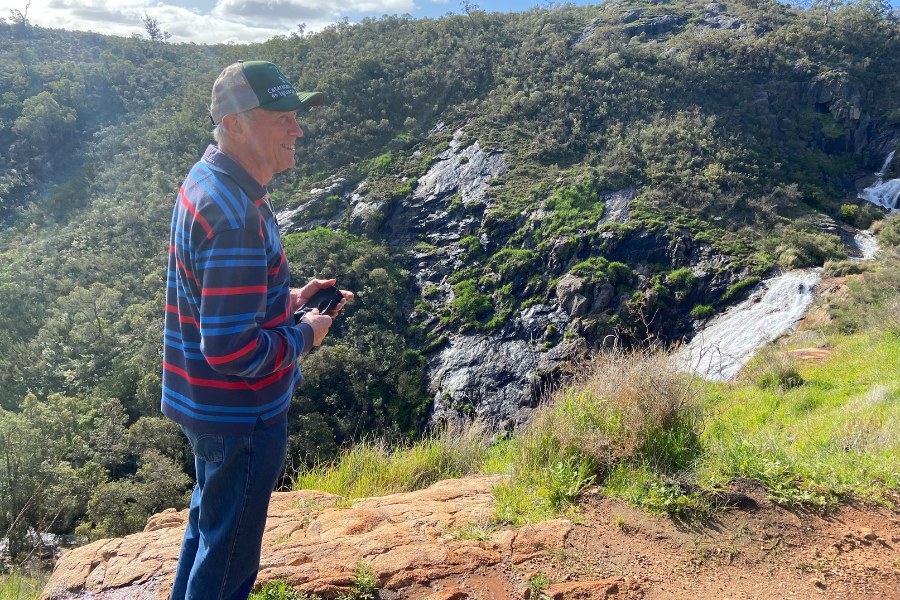 A photograph of Mike Robinson. He is an older man, perhaps in his 60s or 70s. He wears a cap, a long sleeve shirt and jeans. He stands on a trail at Mundy Regional Park that overlooks Lesmurdie Falls. The background is made up of a hill, mostly green with shrubs, but a few dark grey boulders.