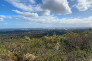 A photo of the view from a very high point at Mundy Regional Park. The view is of the Perth landscape, looking north towards the city. The sky is blue with a few grey clouds, and green bushland takes up the foreground of the photo.