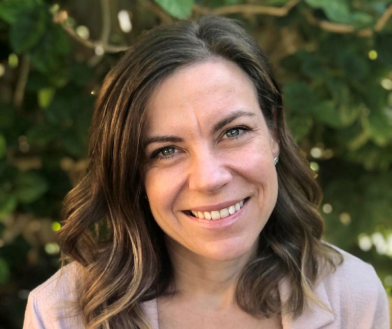 Photograph of Dr Kelsie Prabawa-Sear, CEO of Nature Play WA. Kelsie has light brown, wavy hair, light-skinned complexion and green eyes. Her head and shoulders are visible in this photograph. She is wearing a light pink coloured jacket and is in front of a dark green leafy background.