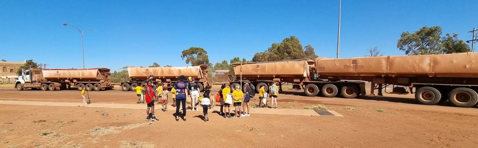 Photograph of Cue Primary School students, doing a Talk N Walk to school. The streetscape shows red dirt beside the asphalt road, with the students and parents in the mid-ground of the photograph, and a multi trailer road train in the background. Behind the road train are large, gum trees and a clear blue sky.