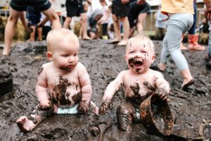 A close-up of two babies sitting in a mud pit. The lower half of their bodies are completely covered in mud, and some mud is splattered on their faces.