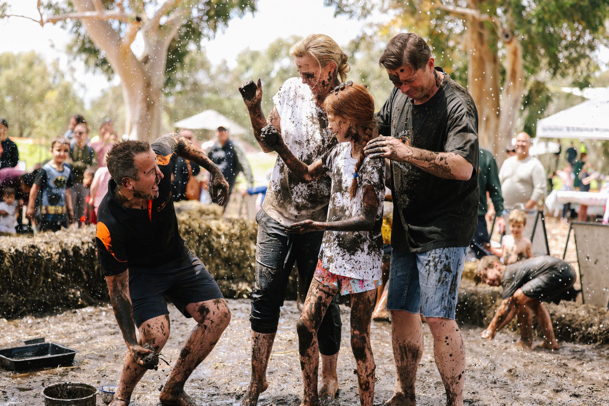 Three adults and one child stand in group in a mud pit. Their clothes are covered in mud. One adult is bending down to scoop up some mud to throw at the others.