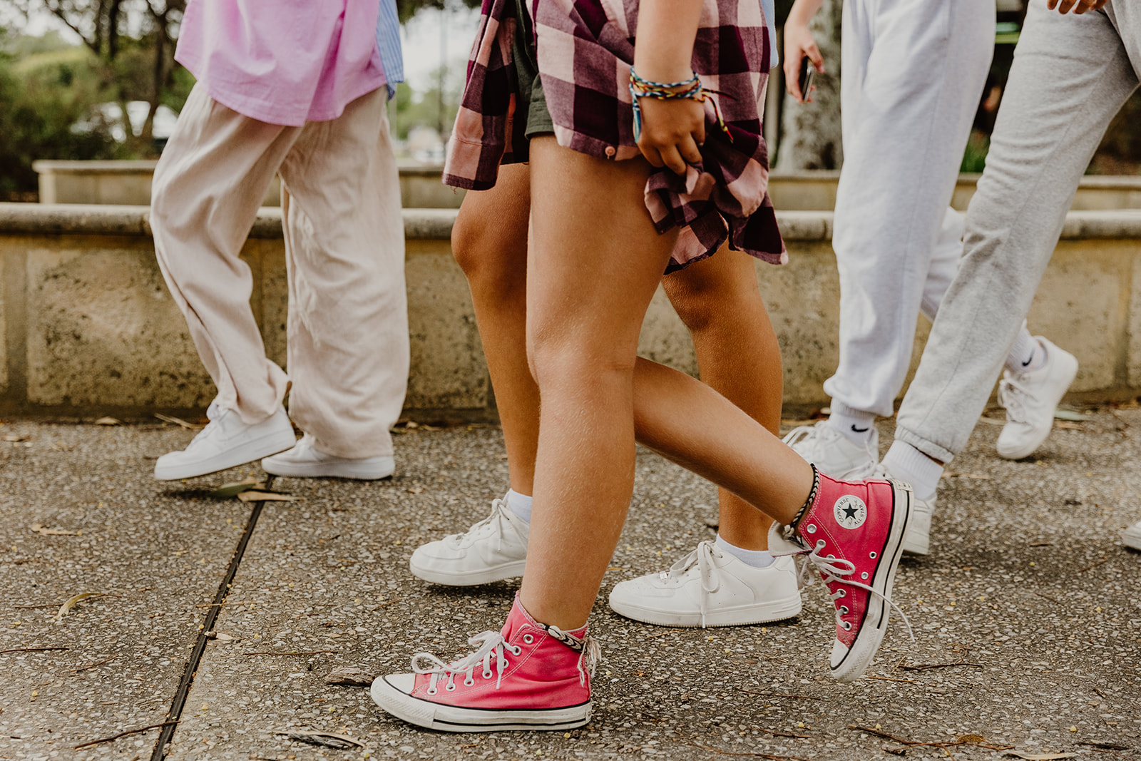 photograph of girls legs, from the waist down, walking to the left of the photograph. From left to right the girls are wearing: girl 1: pink and blue oversized shirt, cream coloured wide-leg pants and white sneakers, girl 2: olive green shorts, pink and maroon checked shirt tied around her waist, hot pink coloured Converse sneakers, and she has friendship bracelets around her left wrist, and one on each ankle, girl 3: light blue jumper, denim shorts and white sneakers, girl 4 and 5: light grey tracksuit pants and white sneakers.
