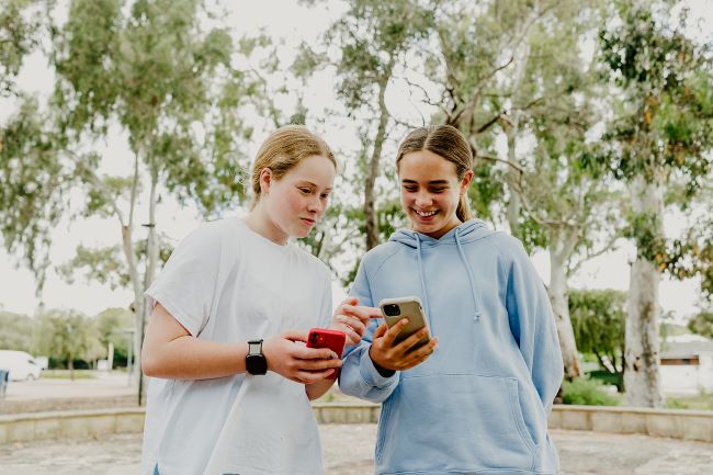 Two girls face the camera, holding the mobile phones and looking at their screens.  The girl on the left is blonde with her hair tied in a low ponytail.  She wears a white t-shirt and a black smartwatch, and is holding a red iPhone.  The girl on the right has brown hair tied back in a low ponytail, and wears a light-blue overshized hoodie.  She holds a gold iPhone.