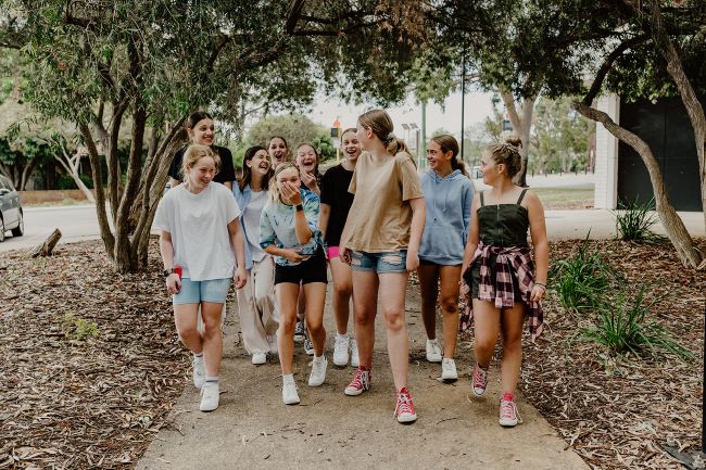 A group of ten girls aged 10 to 13 walking towards camera, on a cement path. They are talking, smiling and laughing as they join in a group conversation. There are tall gum trees along the sides of the path and in the background.