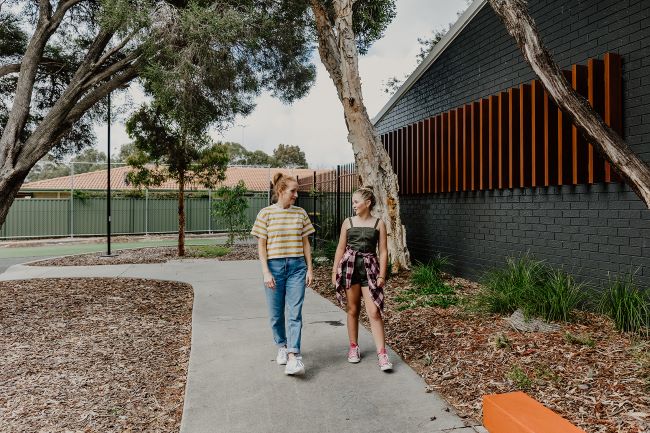 A long shot showing a woman and a girl walking towards camera. They walk along a cement path, next to a dark grey building, and a large gum tree. The woman (on the left) has red hair tied up in a high ponytail, and wears a yellow and white striped t-shirt, blue denim jeans and white sneakers. The girl (on the right) has blonde hair worn in a high bun, and wears an olive strappy playsuit, with a pink and maroon checked shirt tied around her waist and pink high top Converse sneakers. They are talking to each other as they walk.