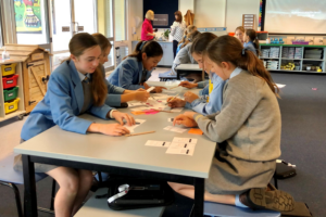 Photograph of a classroom, with a long desk in the foreground, girls sitting either side of the table. The girls wear school uniforms - grey dresses and jumpers with blue blazers and brown leather lace-up shoes. On the table are lots of post it notes with notes written on them.