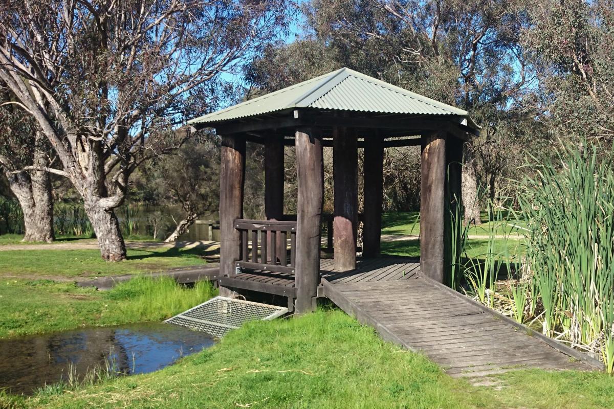 This photo shows a dark wooden gazebo with a light green tin roof. Short wooden boardwalks branch out from three (3) corners of the gazebo. There is water from the river running up to the gazebo. The water is brown in colour and flows through green grass. Long green grass grows to the right of the gazebo, and tall trees surround the area which leads to the brown river. The blue sky can be seen through the tree foliage.