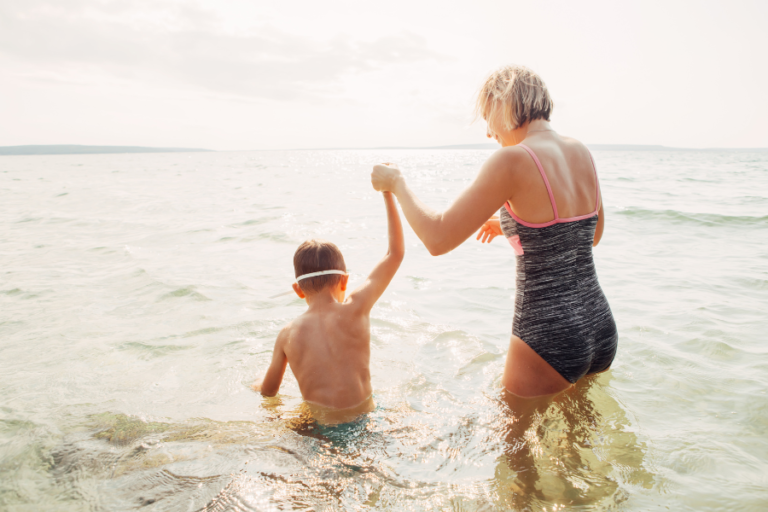 An older woman holding a child's hand as they walk into the ocean.