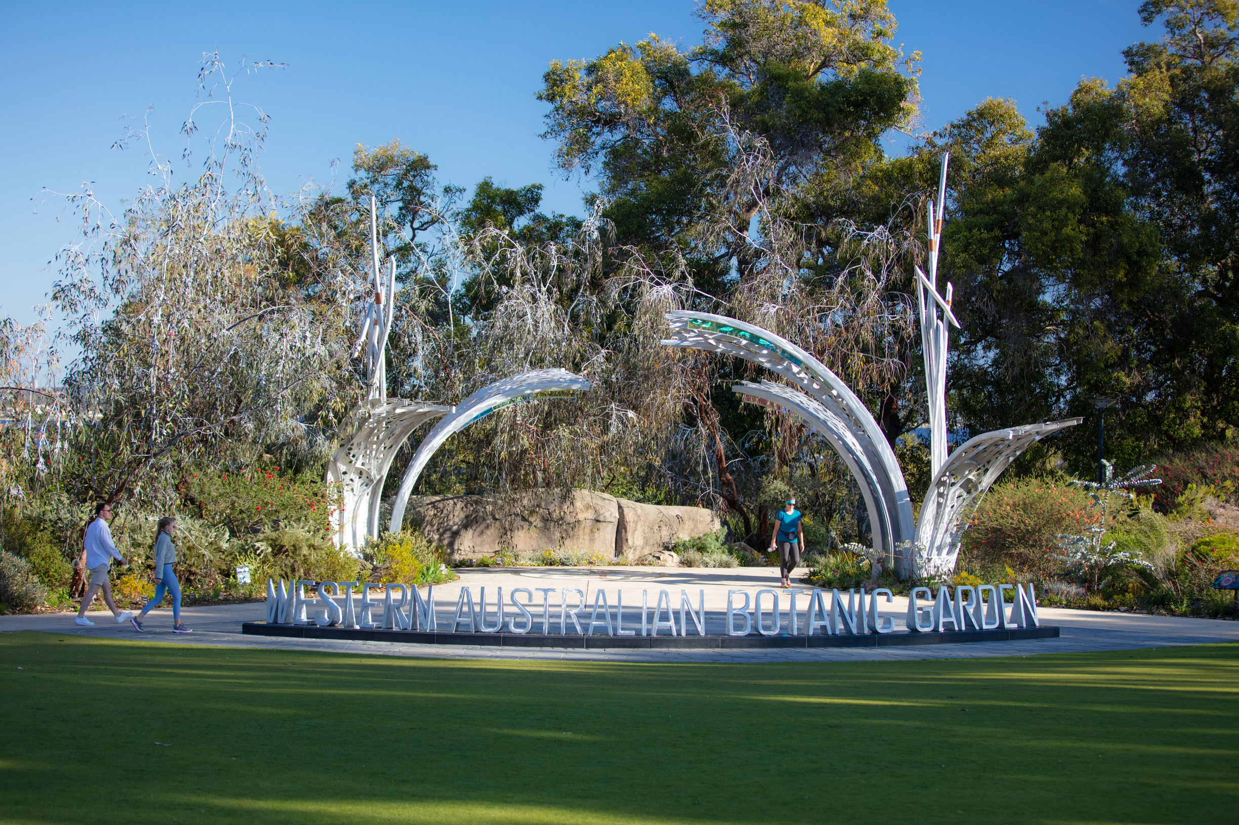 This photograph shows the entrance to the Western Australian Botanic Garden. A grey steel sculpture and sign reading “Western Australian Botanic Garden” in capital letters marks the entrance. There are two (2) people walking from the left towards this sign, with one (1) person walking out of the Botanic Garden. Native plants and trees provide a colourful background with various shades of green and yellow with pops of red. The sky above is blue.