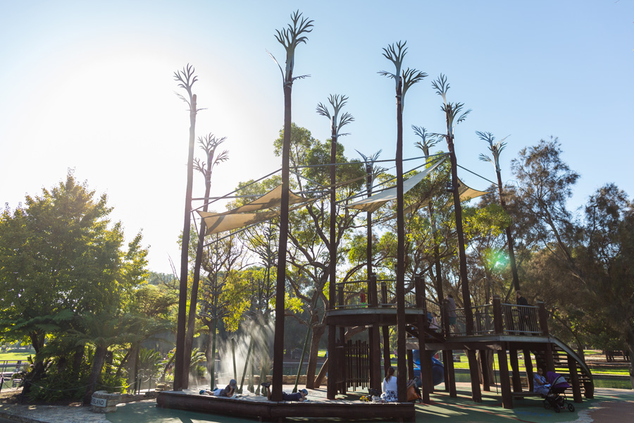 This photograph shows Lycopod Island at May Drive Parkland. The photo shows the replicas of lycopod trees. These are very thin and tall with branches that appear white in colour. A play space with a sandpit, stairs, a slide and a water misting forest sits beneath the trees and is shaded by white shade sails. Green trees and bush surround the play space, where people are resting and playing. The green grass of May Drive Parkland is seen between the play space and trees. The sky above is light blue and appears white on the left due to the glare of the sun. 