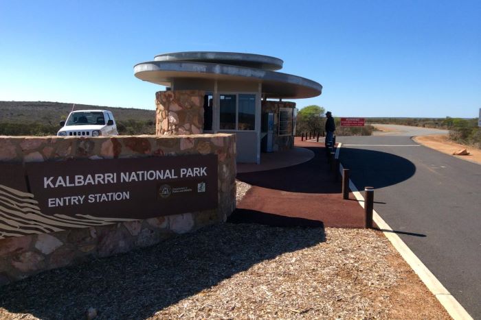 This photograph was taken at the entry station in Kalbarri National Park. The photo shows a sign reading “Kalbarri National Park Entry Station” in white, capital letters. The sign also features two (2) small white logos. A small building is behind the sign with a small glass window and signs. A white car sits to the left of the building. A person stands to the right of the building, where the pathway meets the road. The bushland can be seen in the distance. The sky above is blue.
