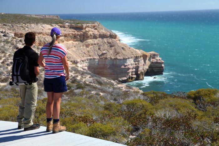 Two (2) hikers stand on the edge of a light grey boardwalk with their backs to the camera. They are looking out to the coastal cliffs and ocean in Kalbarri National Park. The cliff face is cream in colour and drops down into the light blue ocean. Small plants and shrubs surround the pathway and coastline. These plants are mostly green in colour with patches of brown and grey.