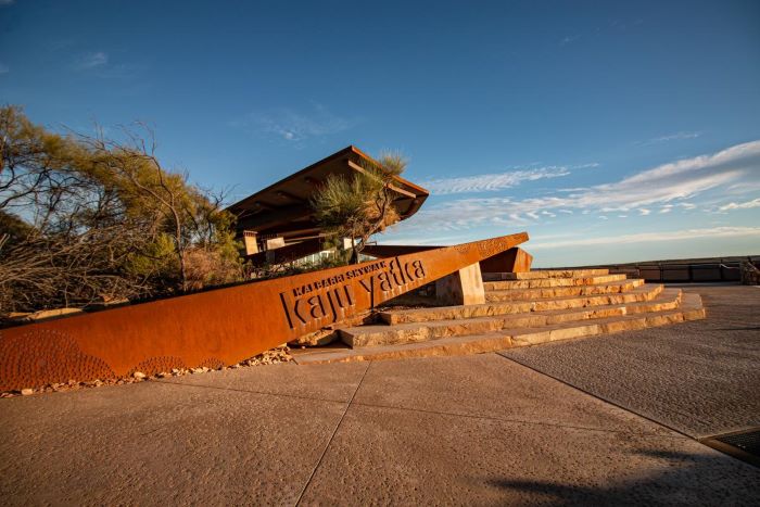 This photograph shows some of the beautiful sculptures and signage at Kalbarri Skywalk. This photo shows the copper-coloured sign reading “Kalbarri Skywalk. Kaju Yatka”. A grey concrete surface leads to stone steps. A building with a square roof sits behind the sign and stairs. Small trees with thick branches and brown and green leaves grow behind the sign. The sky above is mostly blue in colour with streaks of white cloud.