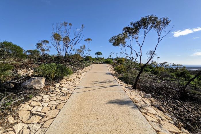 This photo shows the flat concrete path that leads to the top of Meanarra Hill. The path is light grey in colour and features a light cream stone border on both sides. The pathway is surrounded by rocks and bushland that is mostly brown and green in colour. The sky above is mostly blue with a few small white clouds seen on the right of the image.