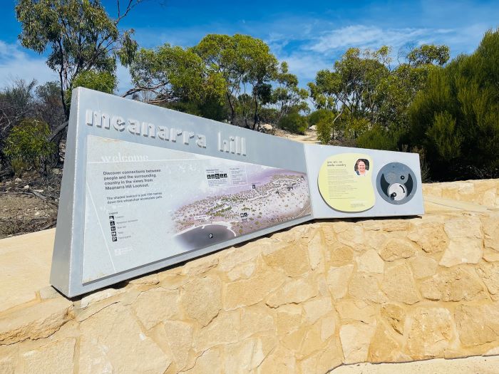 This is a photograph of an interpretive sign at the bottom of Meanarra Hill. The sign is silver in colour and reads “meanarra hill” in embossed lettering and features an illustration of the walk path with information and icons, as well as a Welcome to Country by a local Nanda representative. The sign is attached to a cream stone wall. The bush environment grows behind the sign with shrubs and small trees with green leaves. The sky is seen above and is mostly a deep blue colour with patches of white fluffy clouds.