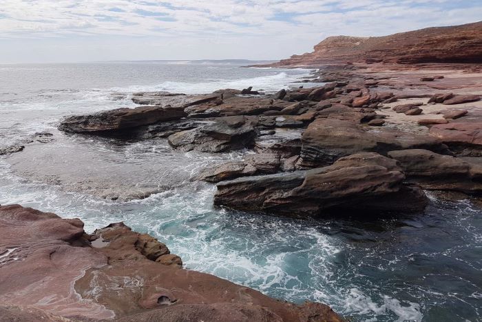 This photograph shows the rocky waters near Mushroom Rock. The rocks are mostly red and brown in colour and lead to the blue ocean. The sky above is mostly white with fluffy clouds. A red cliff face stands on the right of the image and is mostly red in colour.