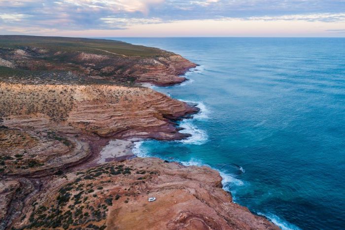 This photo was taken using a drone and shows the rocky coastline in Kalbarri National Park. This location, known as Pot Alley, features rough and curvy cliffs that are brown/red in colour with green plants covering the landscape. A small beach with white sand is located between two (2) cliffs. The ocean crashes onto the rocks, creating waves that appear to be white. The ocean is mostly light blue in colour with patches of darker blue. The sky above is cloudy and is a deep grey colour with patches of lilac, pink and blue. A white vehicle can be seen towards the bottom of the image.