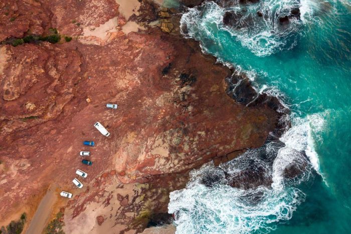 This photograph was taken overhead using a drone, and shows the red, brown and cream rocky cliffs at Red Bluff Beach. There are eight (8) cars parked on the rocky surface which extends down into the light blue/turquoise ocean. The water crashes onto the rocky surface creating white waves.