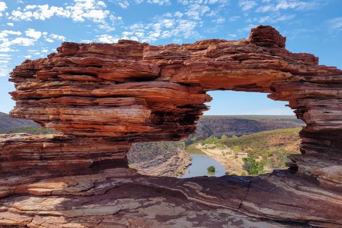 This photograph was taken at Nature’s Window and shows the rock formation that forms an archway and frame. The rocks are a mixture of colours including red, cream and brown. The river can be seen in the centre and is brown in colour. The sky above is blue with patches of white fluffy clouds.