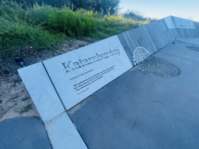 A photo of a long, low interpretive sign, made of concrete and timber. It reads "Katamburdup. A place of high importance. My great-great-great grandmother was taken from around here, her name was Jili and her totem was the white-necked heron - the missionaries called her Matilda. Trevor Walley, 2020".