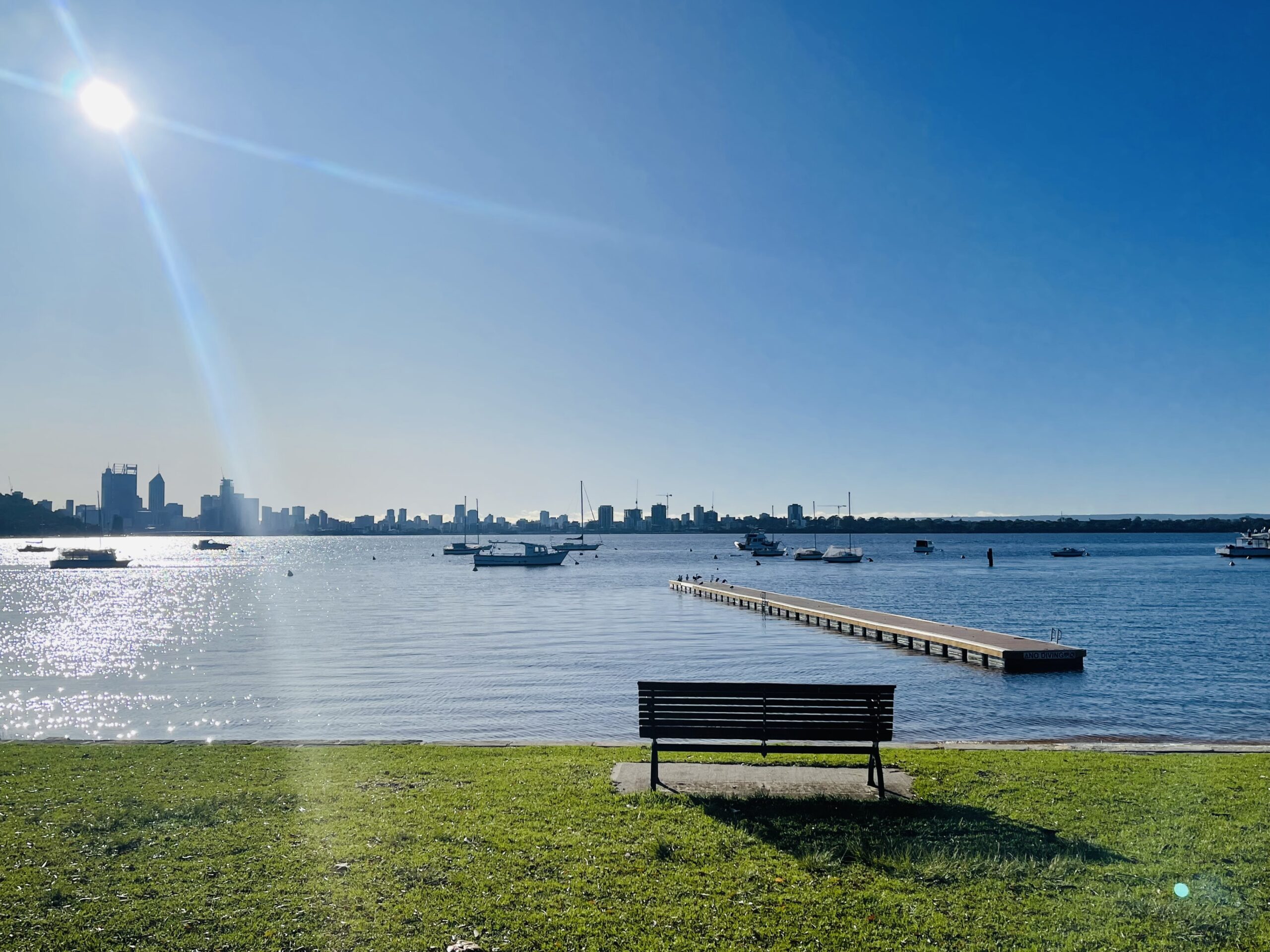 A photo of the Swan River taken from Matilda Bay Reserve. Green grass and a park bench can be seen in the foreground of the image. The midground is made up of the river, on which float a pontoon and some boats. The clear blue sky is above, and some buildings can be seen in the distance.