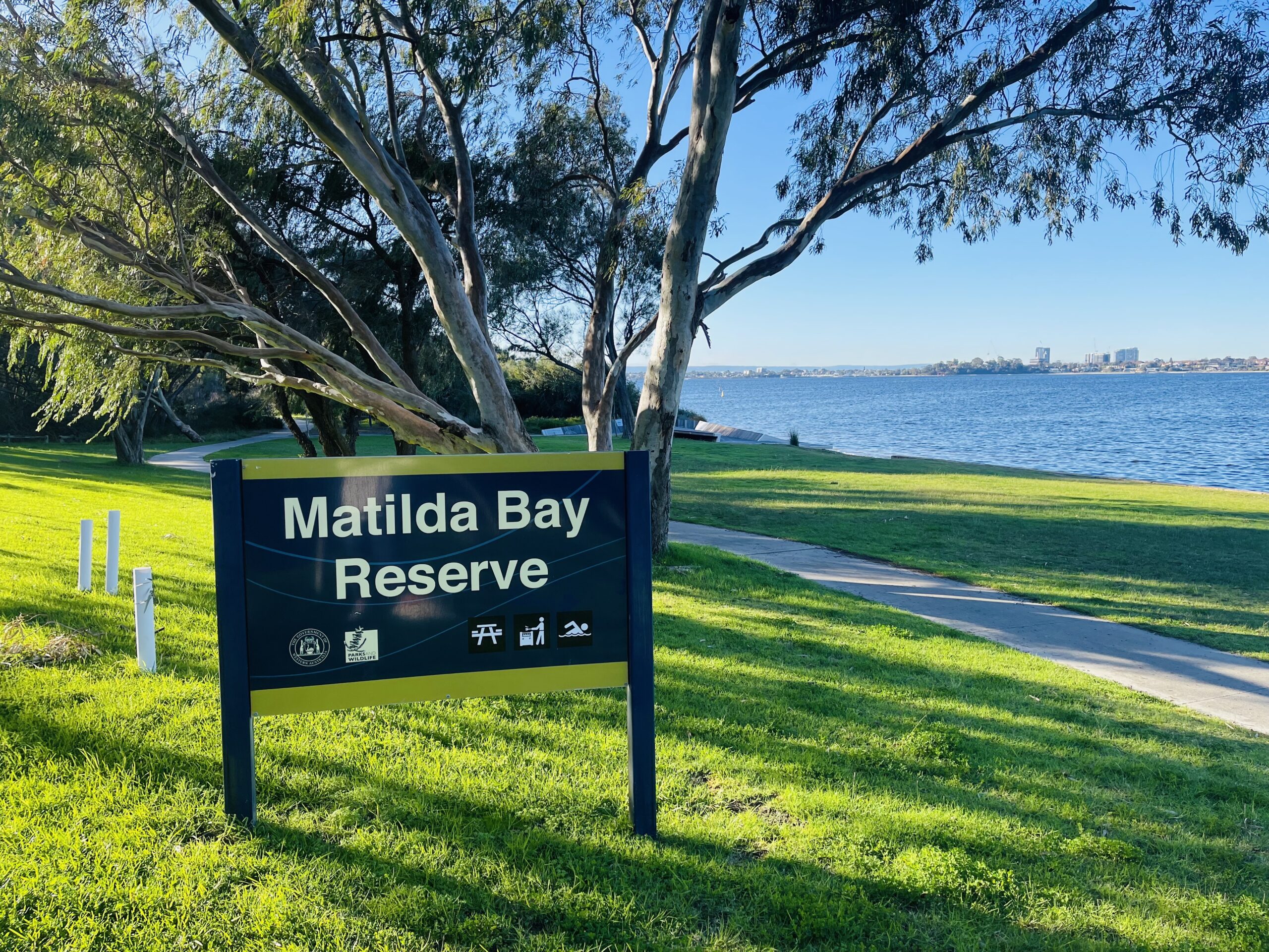 The focus of this photo is a large sign that reads “Matilda Bay Reserve” in the left of the image. Behind and to the right of the sign is some green grass, and a concrete path. Some large trees and the blue Swan River can be seen in the distance. The sky above is clear and blue.