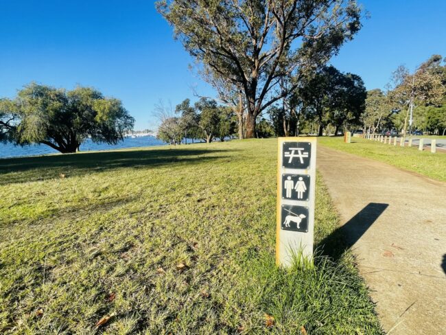 The focus of this photo is a small sign with three icons – a picnic table icon, a male and female toilet icon, and a “dogs on lead” icon. The background of the photo consists of green grass, trees, and a concrete path. The river can be seen in the distance, and the sky above is clear and bright blue.