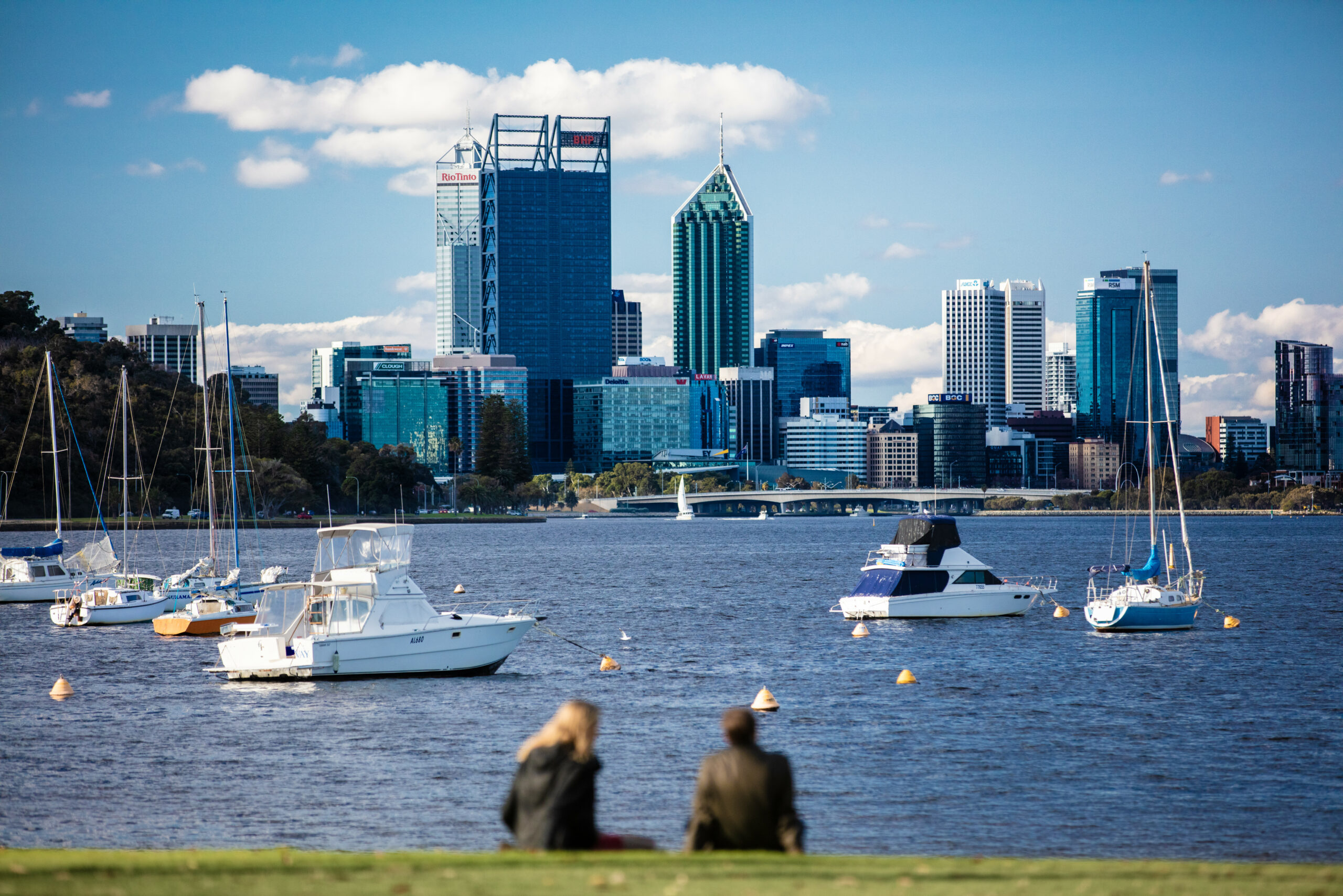A photo of the views from Matilda Bay Reserve. The foreground is out of focus, and features two people sitting on the bank of the Swan River. The rest of the photo is in focus, and features the blue river and some boats floating atop it, the Perth City skyline, and the blue sky above.