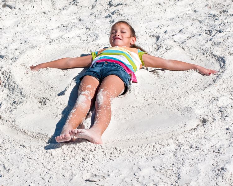 A photo of a child in a t-shirt and shorts, laying on their back on the sand. Their arms are stretched out to the sides, while their legs remain together. Patterns in the sand show the child has been moving their legs and arms in and out, to form the shape of an angel in the sand.