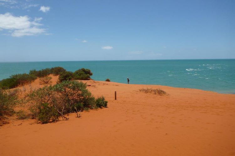 A photo taken at Cape Peron from the top of a sand dune. The rich, orange-coloured sand dune can be seen in the foreground, with a person standing atop it in the distance. In the midground, past the dune, is the turquoise-coloured ocean. Above, the sky is blue and clear, besides a few small white clouds.