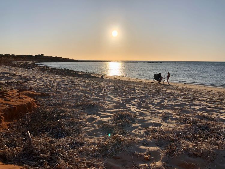 A photo taken at a beach in Francois Peron National Park. In the distance, one person sits in a camping chair, and a child stands next to them holding a fishing rod. They are right by the water’s edge. The water is calm and flat. The sky is clear and the sun is low to the horizon, indicating it’s later in the day.