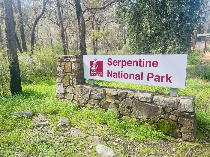 A photo of a sign on a short stone wall surrounded by green grass, bushes and trees. The sign reads “Serpentine National Park.”